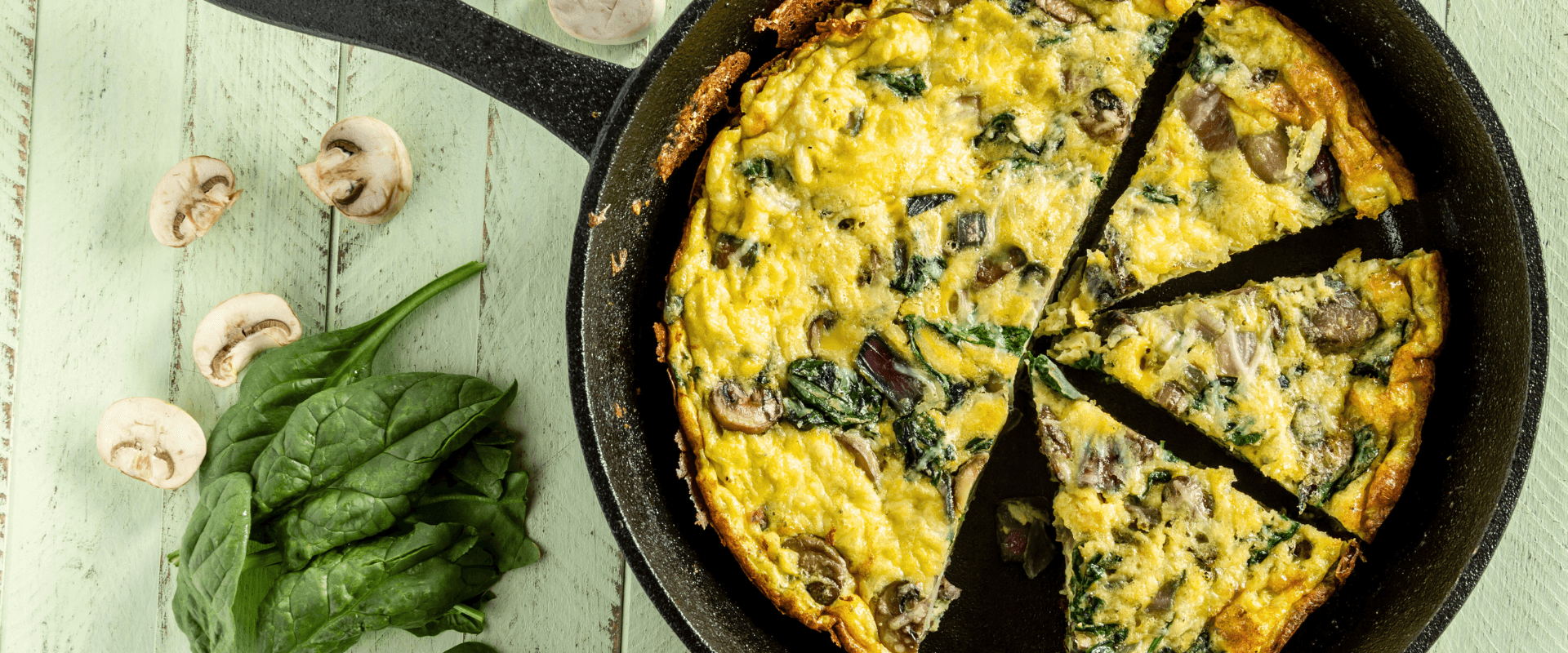 A sliced frittata with spinach and mushrooms in a cast iron skillet, next to fresh spinach and sliced mushrooms.