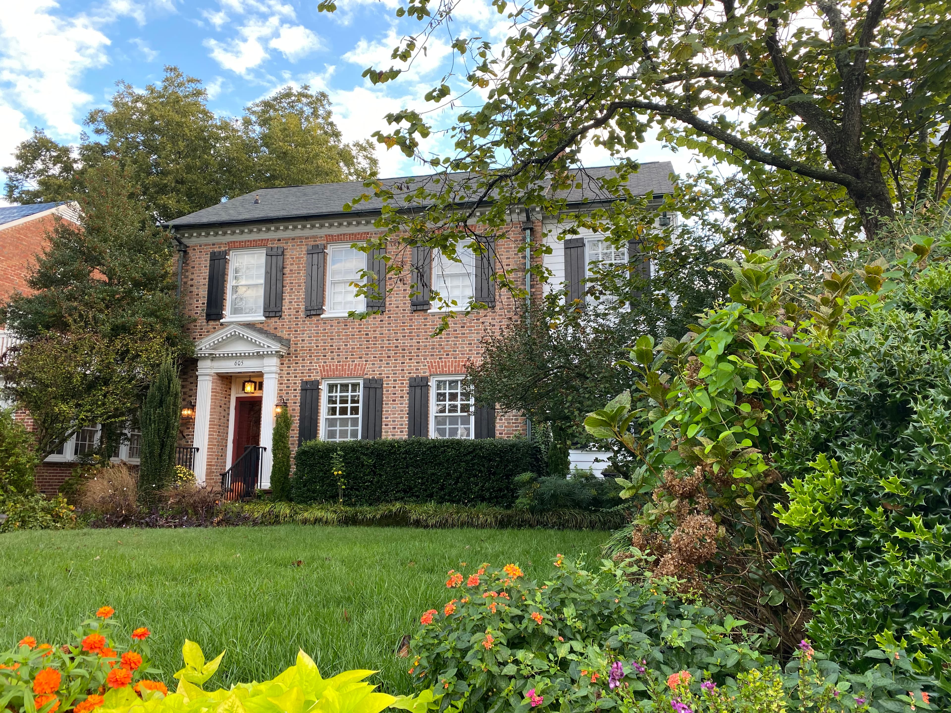 A brick two-story house surrounded by green foliage and colorful flowers.