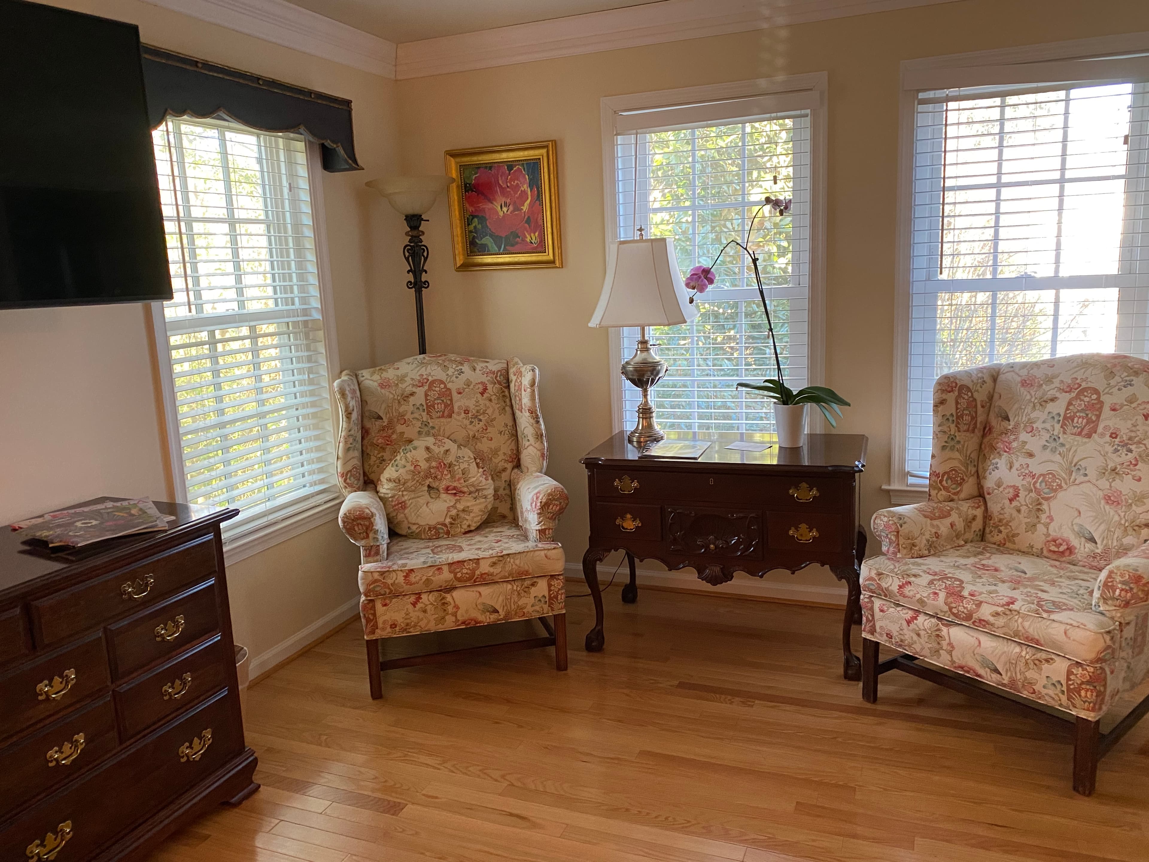 A cozy living room corner featuring two floral armchairs, a wooden side table, a lamp, and a window with sunlight streaming in.