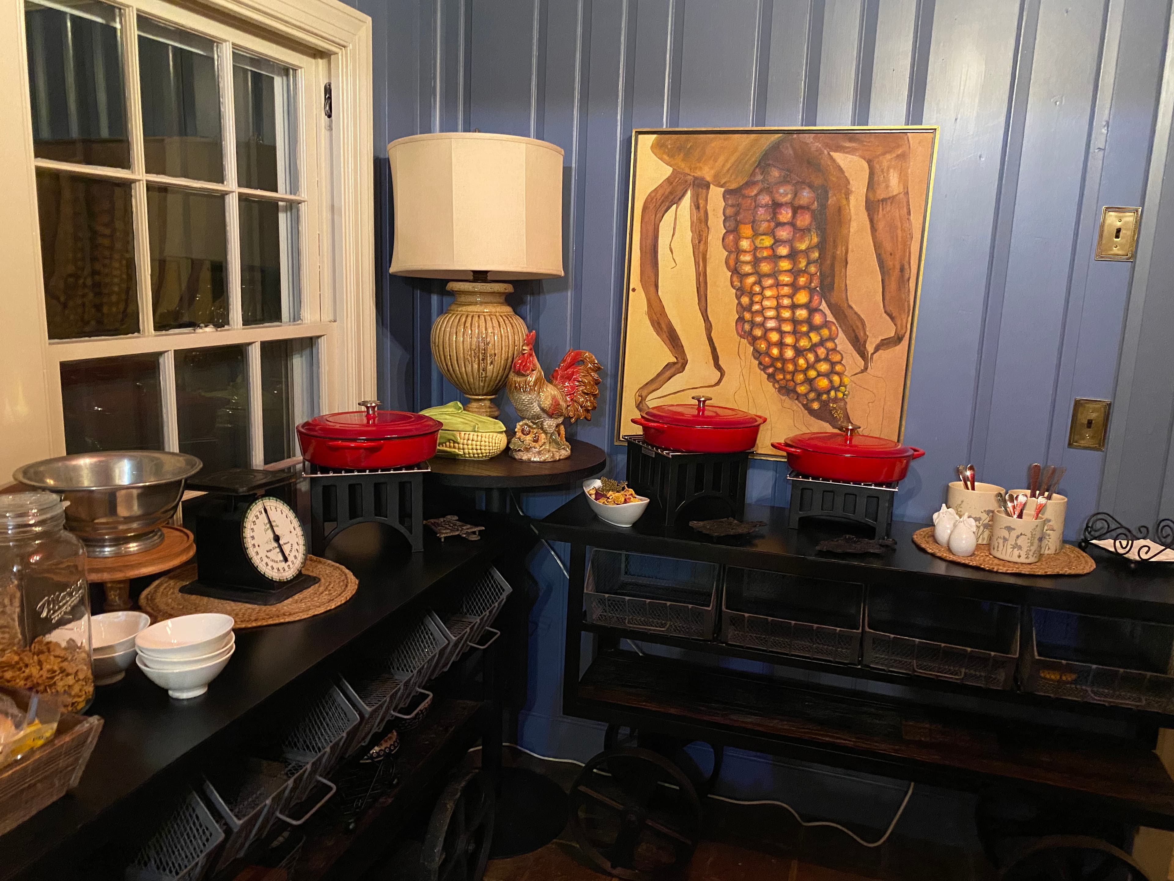 A cozy dining corner featuring red cookware, a lamp, and a corn-themed painting against blue wooden walls.