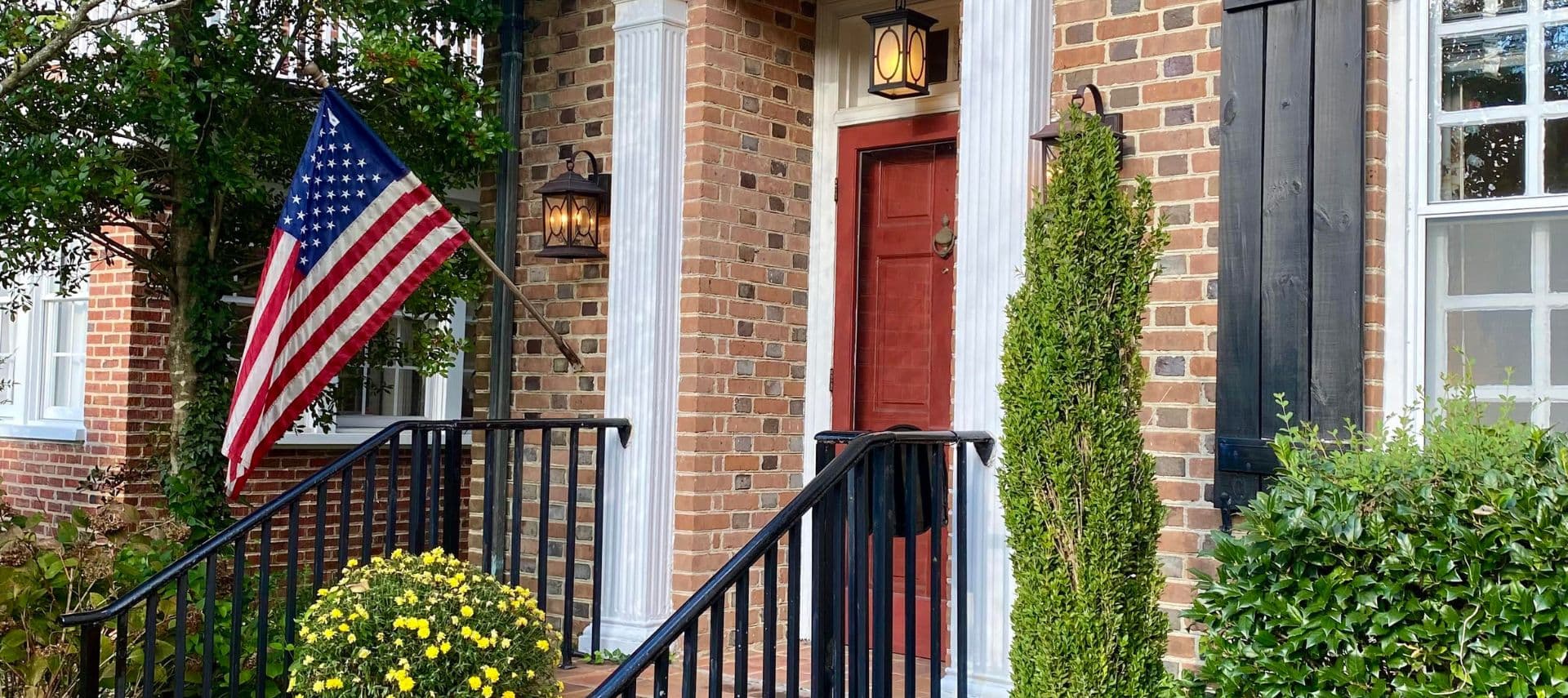 A brick-front home with a red door, flanked by an American flag and greenery.