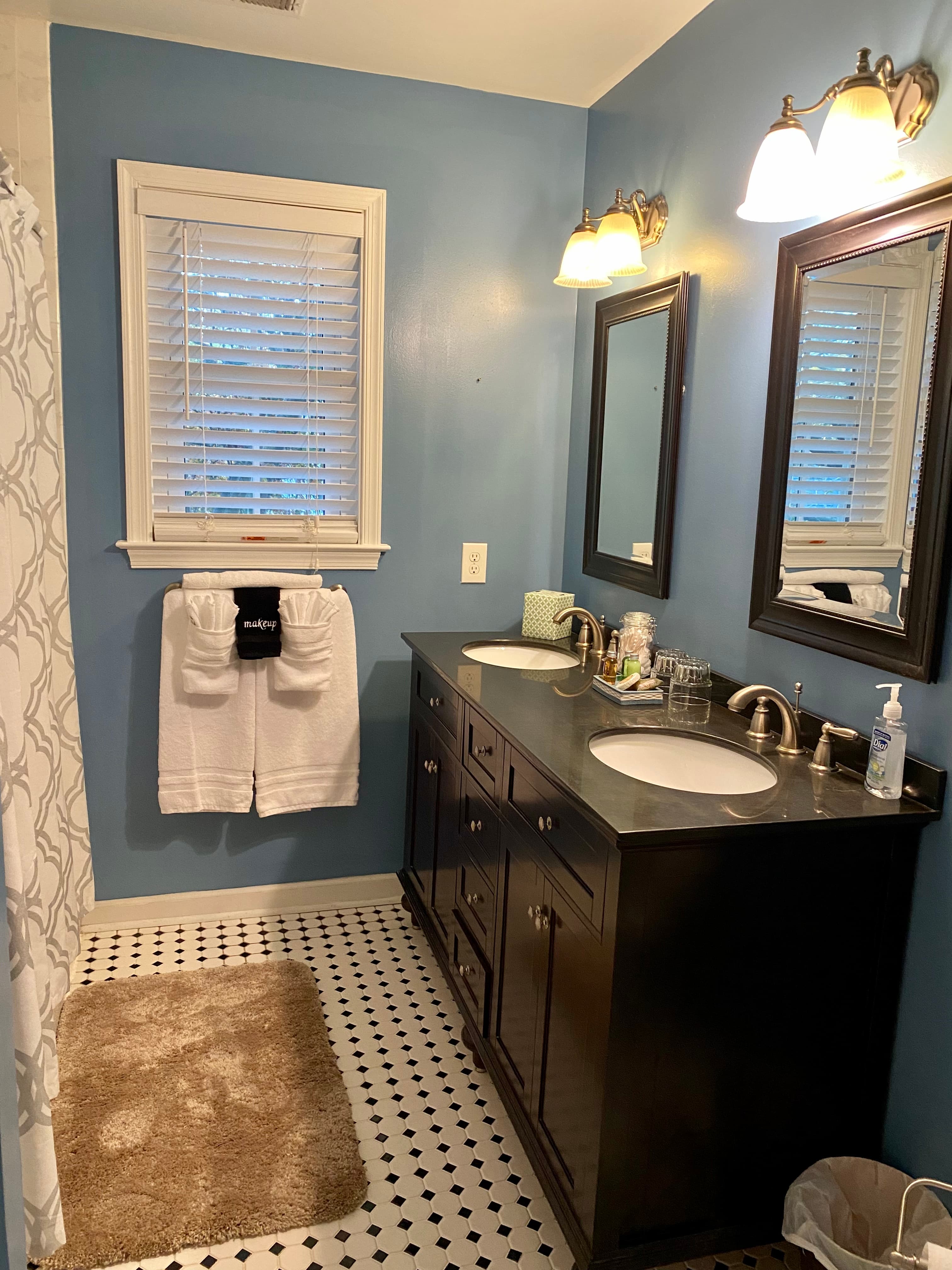 A modern bathroom with blue walls, dual sinks, mirrors, and white towels hanging by a window.