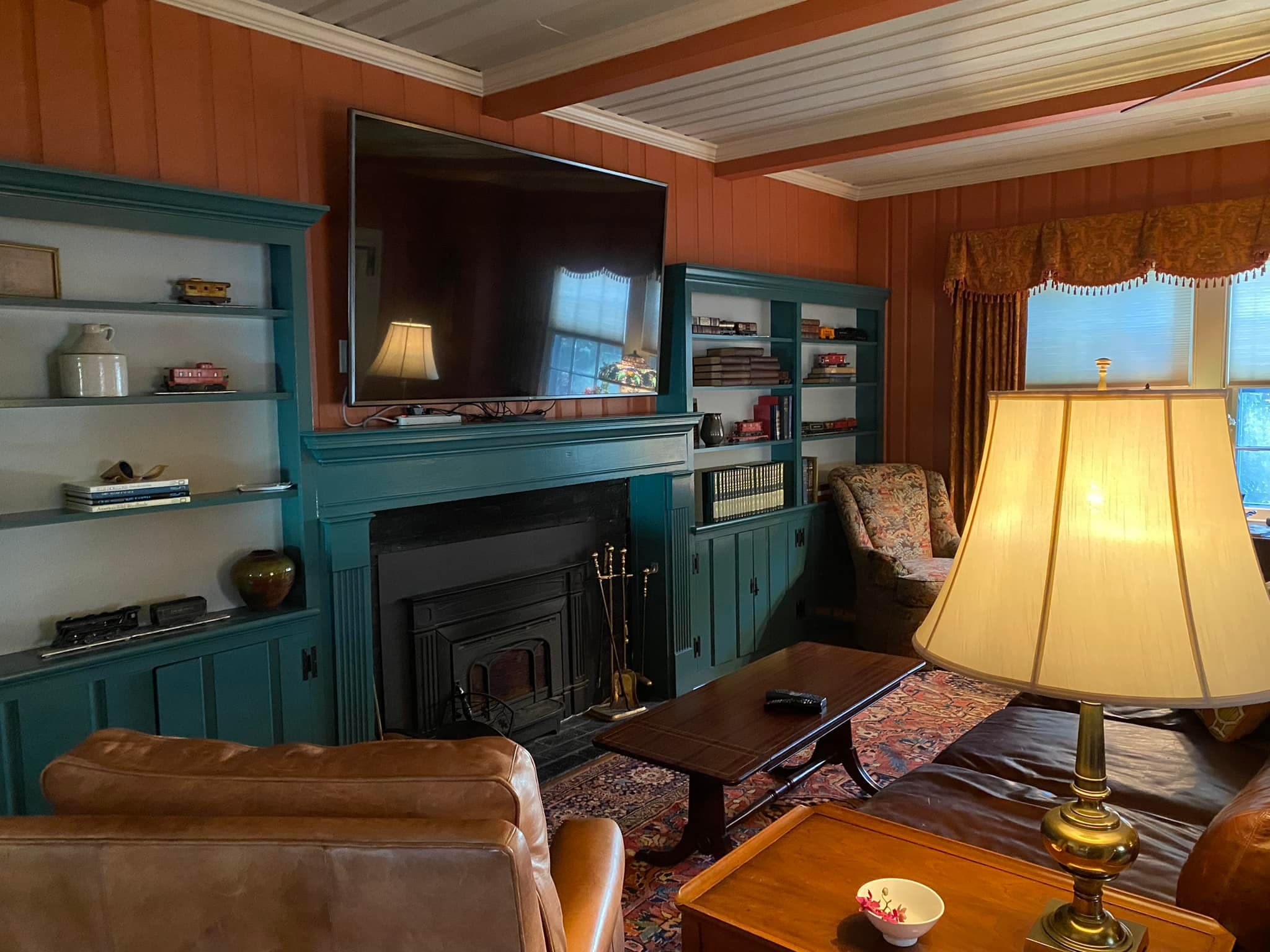 Cozy living room featuring a television above a fireplace, surrounded by bookshelves, a lamp, and comfortable seating.