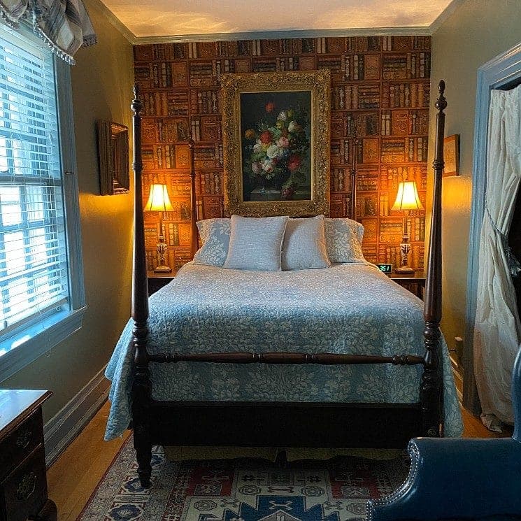 Cozy bedroom featuring a four-poster bed, decorative lamps, and a wall adorned with a book-patterned wallpaper and a floral painting.