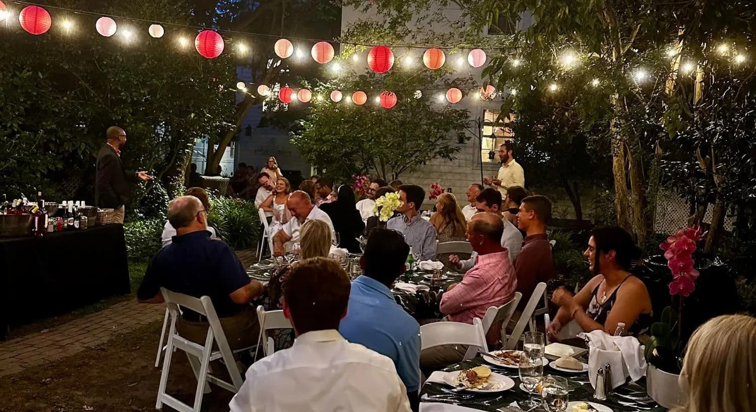 A group of people dining outdoors under string lights and lanterns, attentively listening to a speaker.