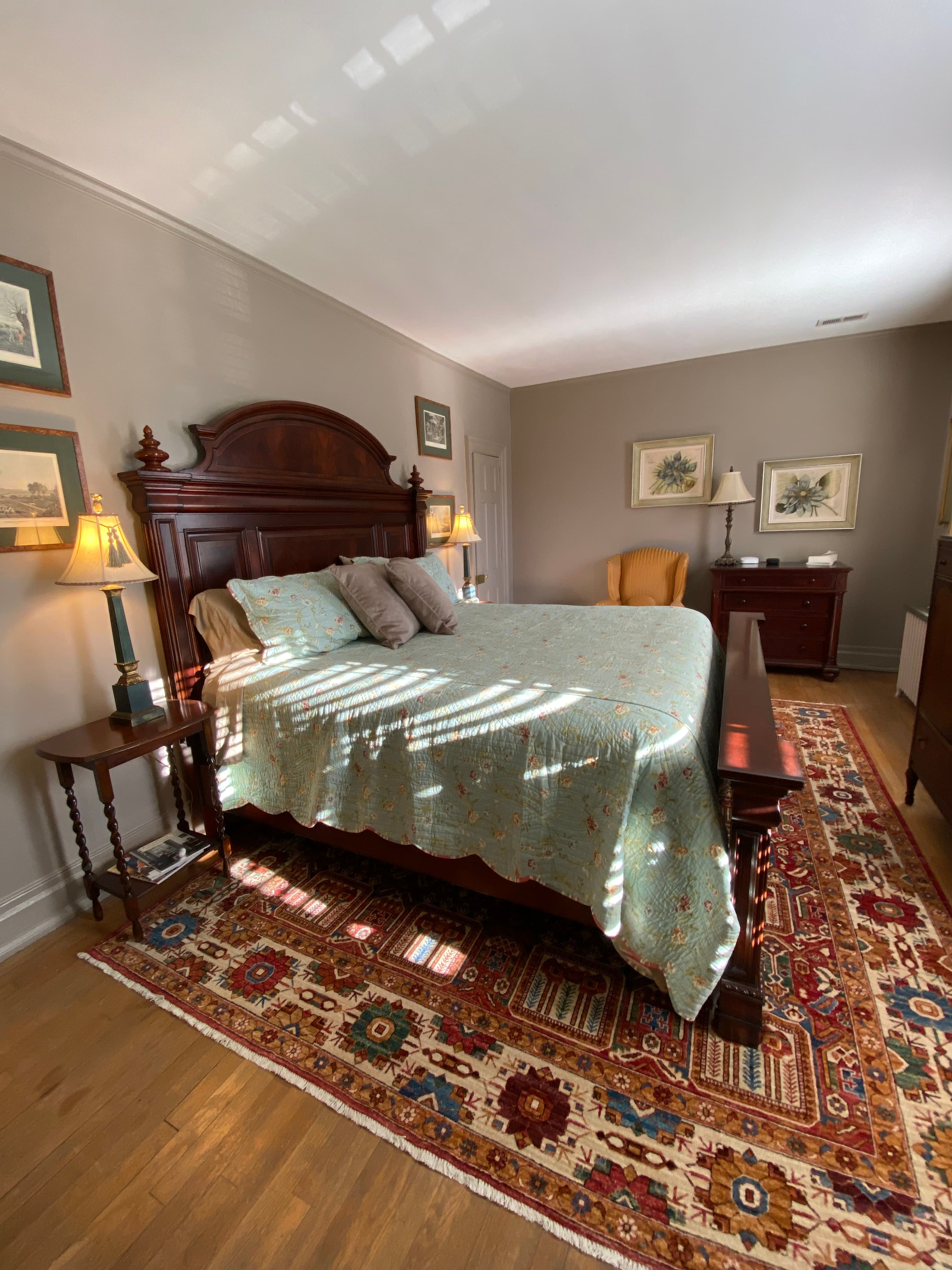A cozy, well-lit bedroom featuring a wooden bed with a quilted cover, a nightstand with a lamp, and an armchair on a patterned rug.