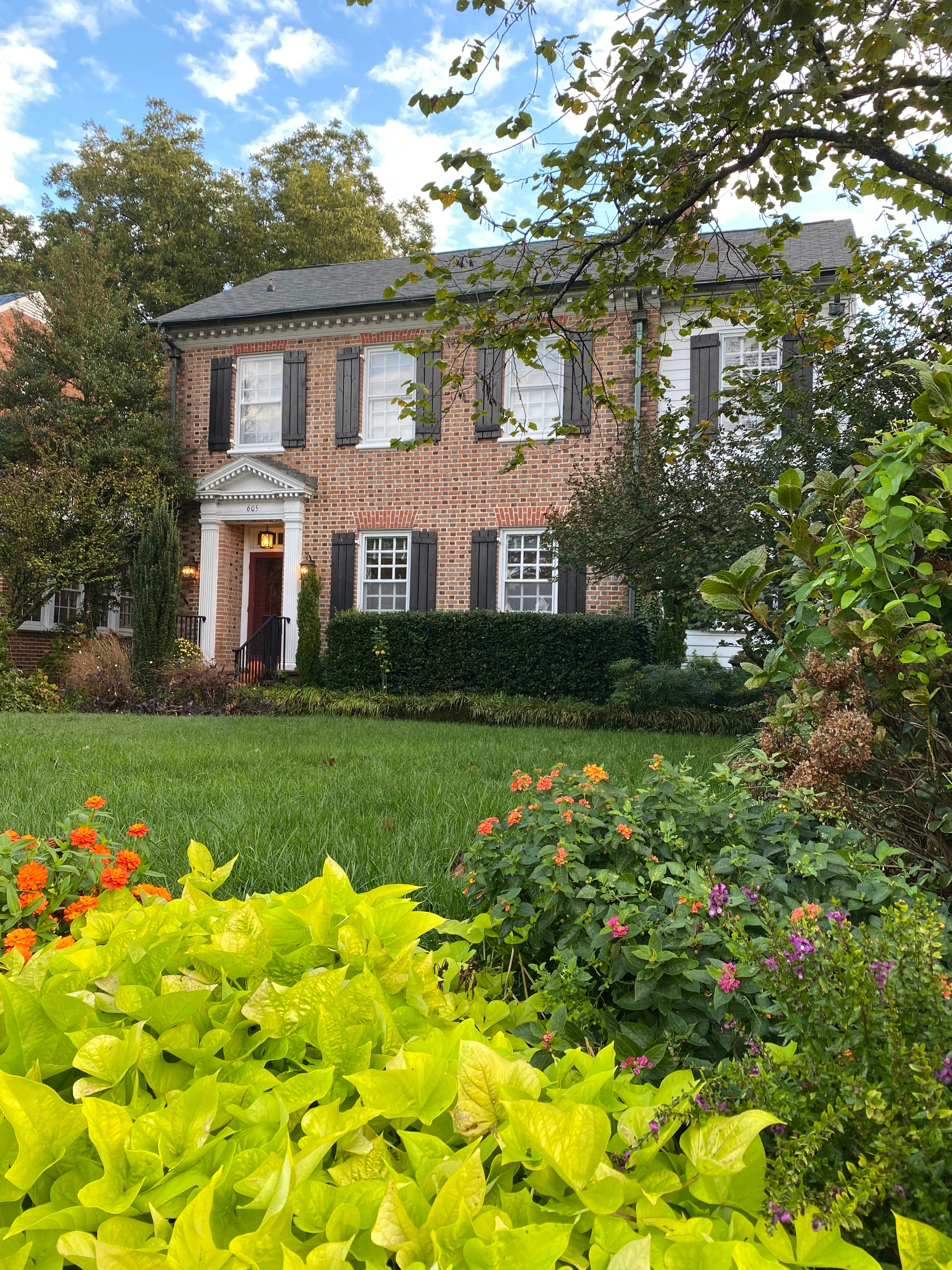 A brick house with black shutters surrounded by colorful flowers and greenery.