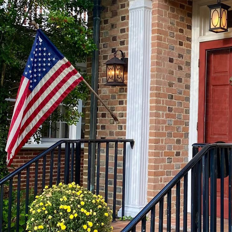 A U.S. flag hangs beside a front door supported by a brick wall with a potted yellow flower.
