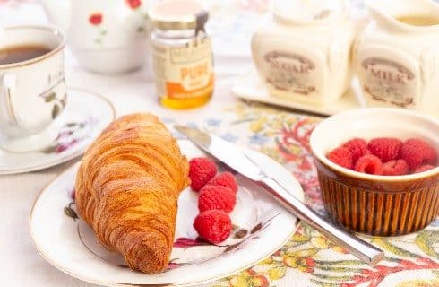 A golden croissant and raspberries on a floral plate next to a jar of honey and cream and sugar