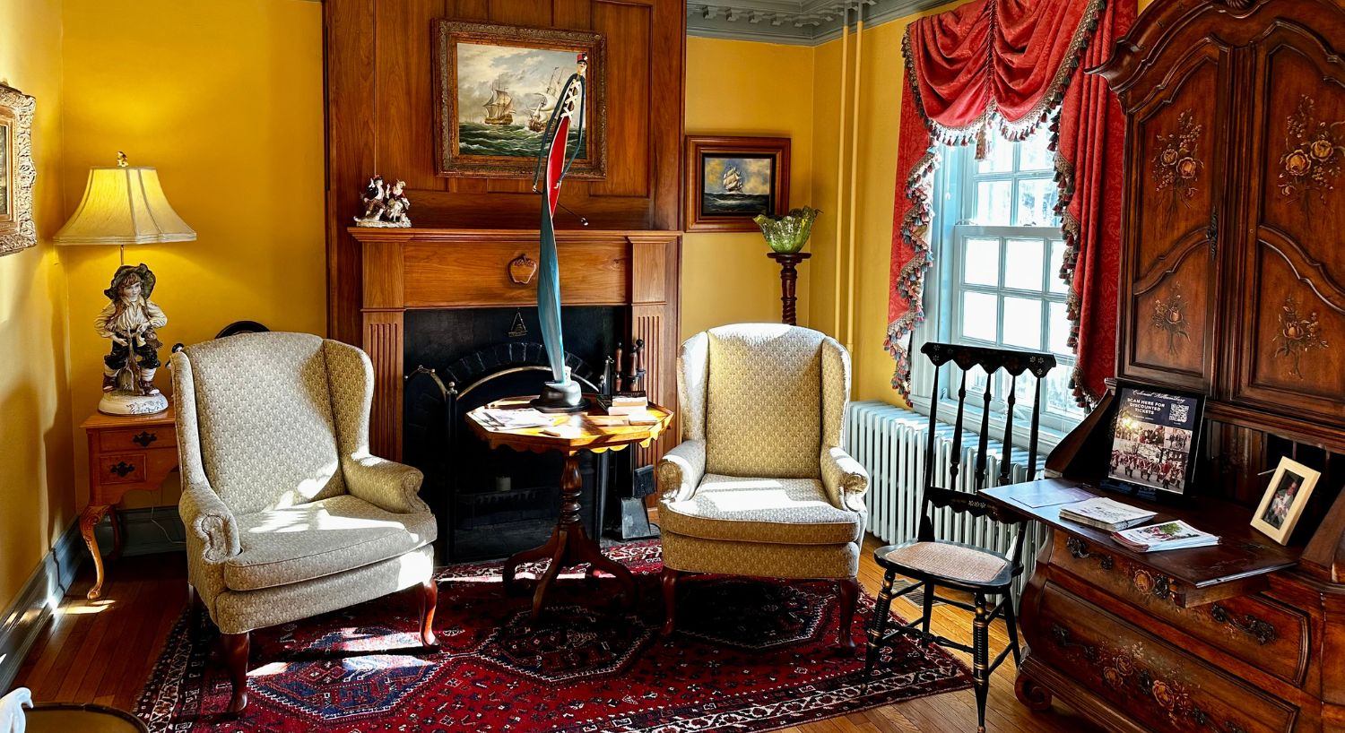 Gorgeous sitting room with two wingback chairs and table in front of a fireplace, yellow walls, window with a red valence draped at the top and large antique hutch