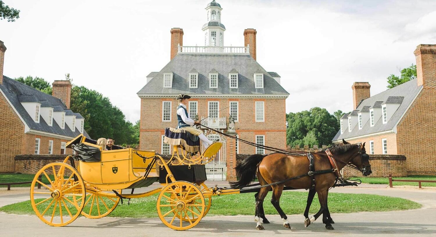 A woman in a yellow antique carriage being pulled by two horses in front of a Colonial estate.