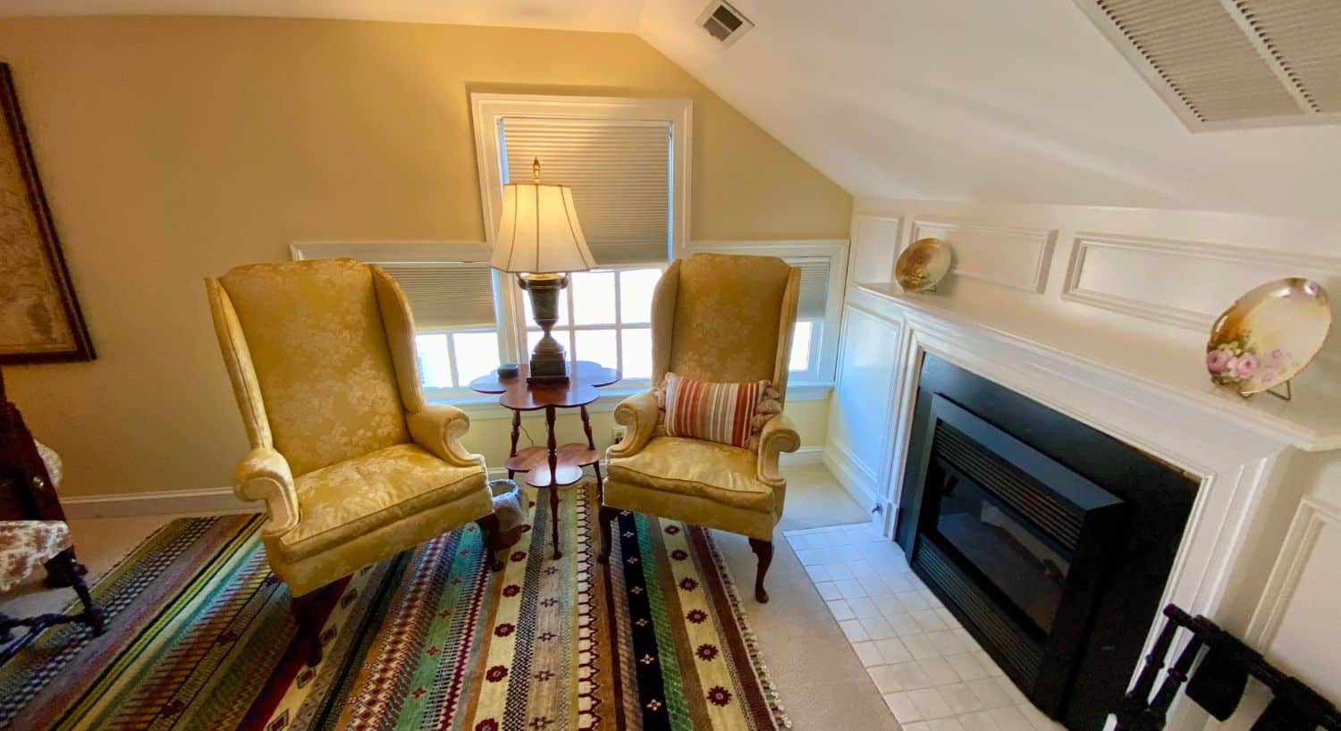 Two gold wingback sitting chairs and a table with a lamp in front of a window and next to a gas fireplace