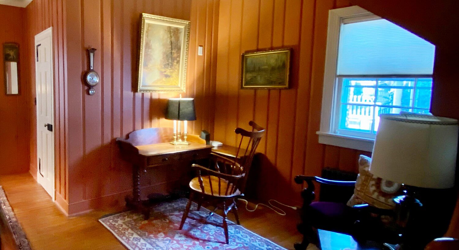 Corner nook of home with wood paneled walls, a writing desk with chair and lamp and one window with blinds