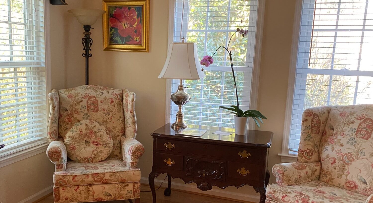 Two floral wingback sitting chairs and an antique table with a lamp and potted flower in front of three large windows