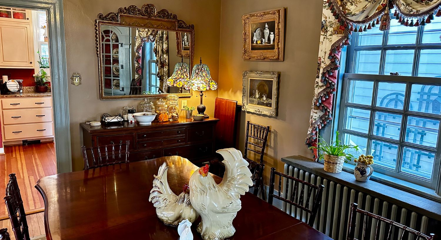 Elegant dining room with antique table and chairs, buffet with serving dishes and jars, Tiffany lamp and entryway into a kitchen