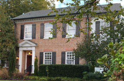 Exterior of a large brick two-story home with many windows with black shutters, large trees and shrubs in front