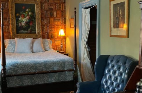 Elegant bedroom with four poster bed, leather wingback chair, side tables with lamps and doorway into a bathroom