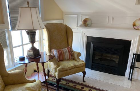 Sitting area of a bedroom with two gold wingback chairs and table with lamp next to a gas fireplace