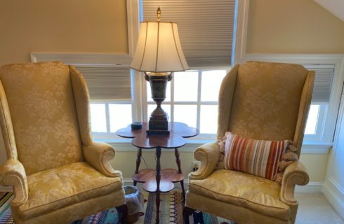 Two gold wingback sitting chairs with a table and lamp near windows with blinds