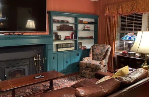 Living room with brown leather couch and floral chair in front of a fireplace with TV above and flanked with built in book shelves