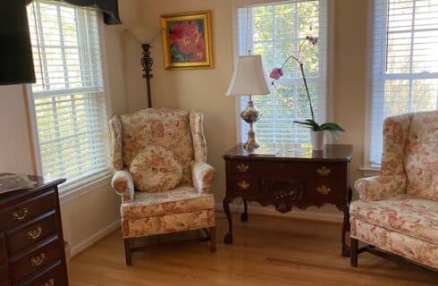 Sitting area in a bedroom with two floral wingback chairs, antique table with lamp and potted flower by three large windows