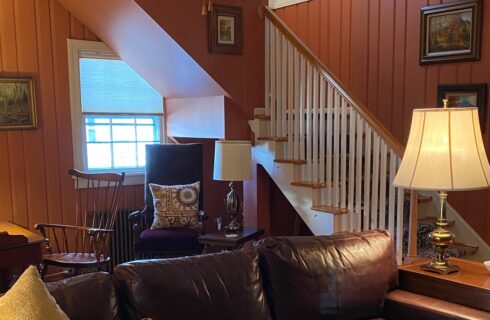 Living room with brown leather couch, table with lamp, nook with writing desk and stairwell to an upper level