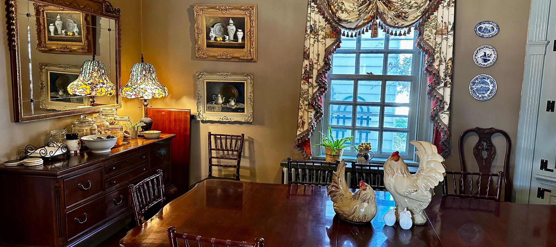 Elegant dining room with large antique table and chairs, buffer with plates and serving dishes and window with floral curtains