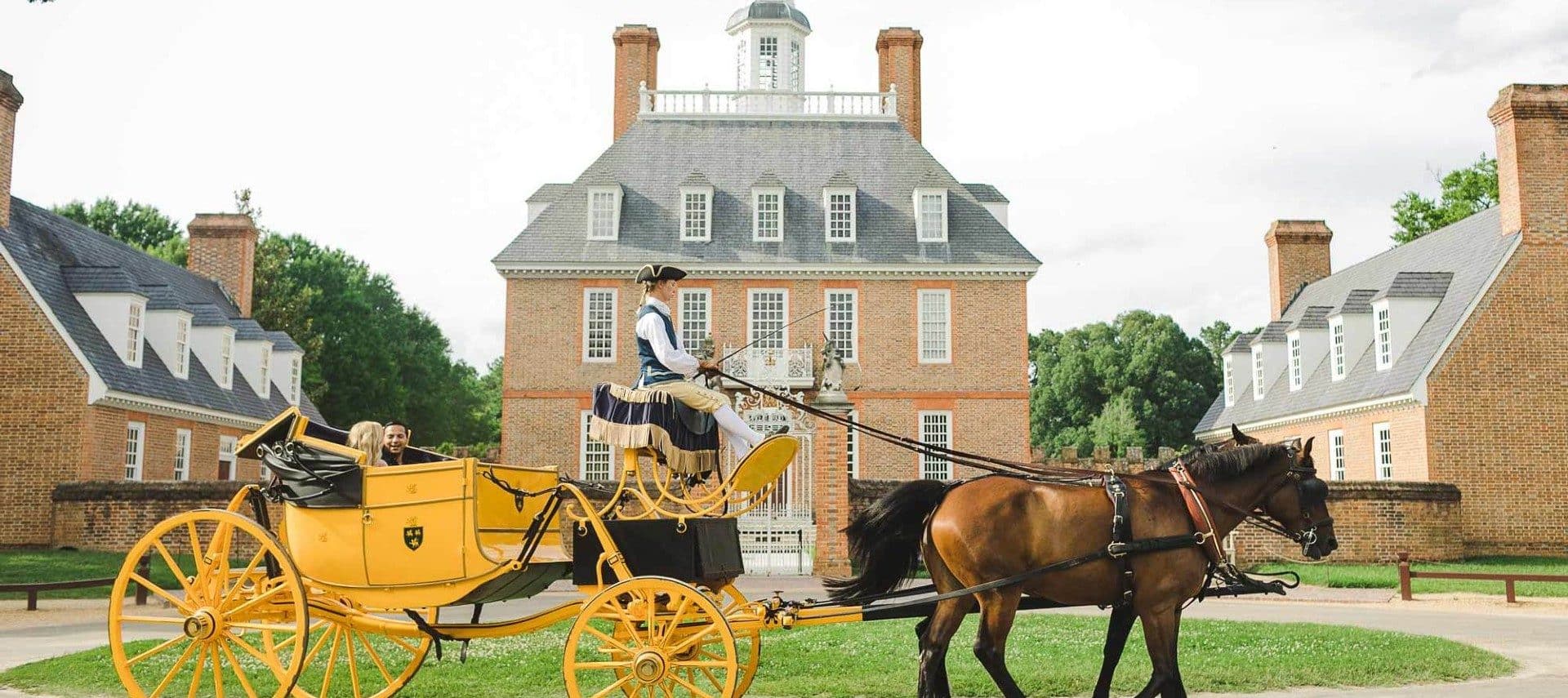 A woman in an antique carriage being pulled by two horses in front of a colonial set of homes