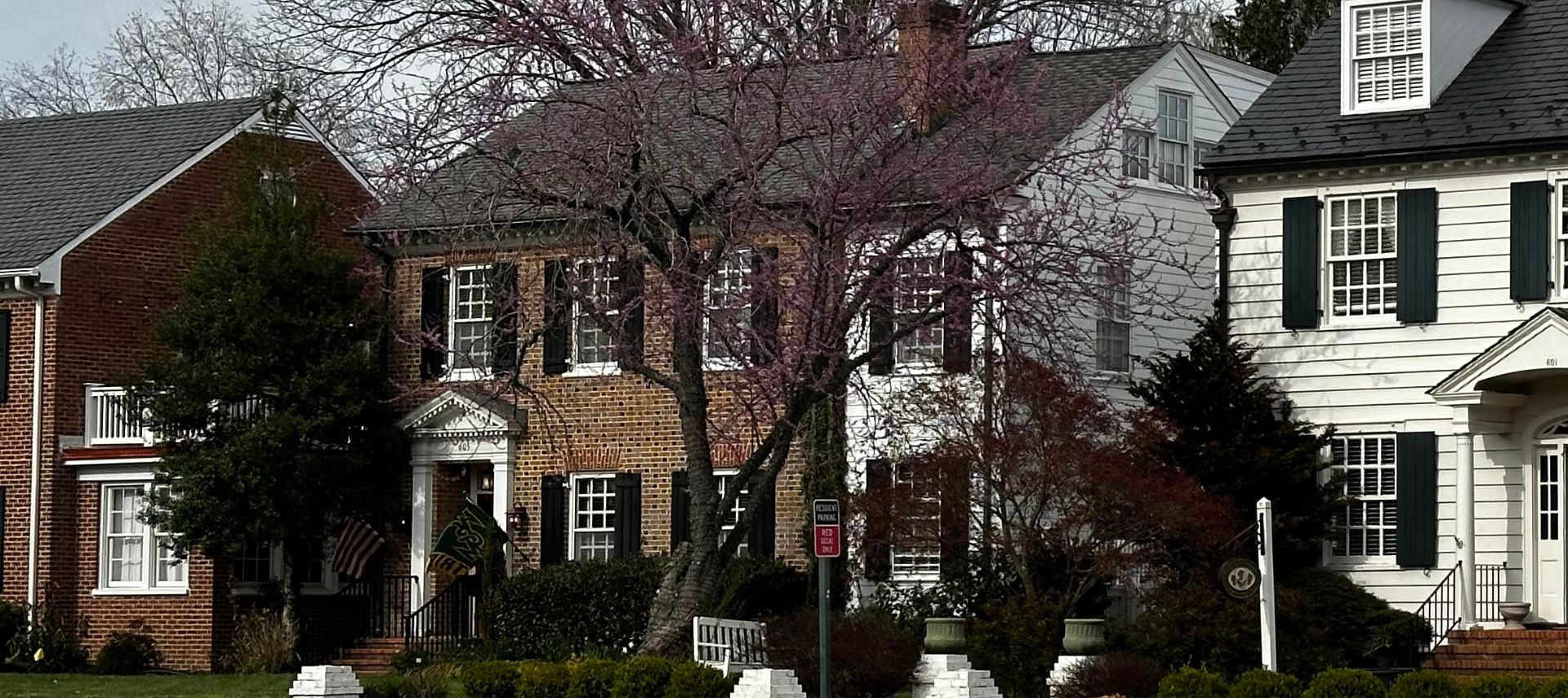 Exterior view of a large, stately brick home with several windows with black shutters, flanked by other homes and large trees in the front yard.