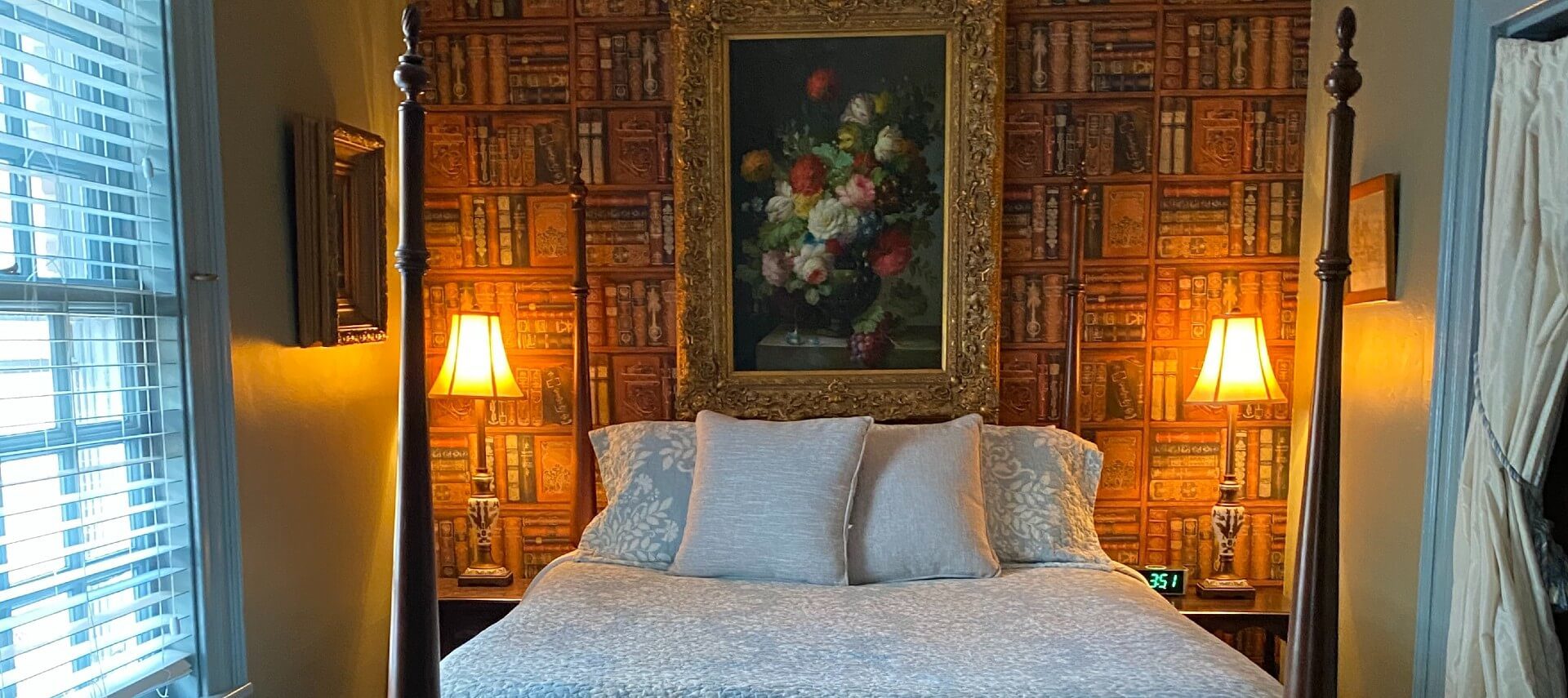 Bedroom with four poster bed against a wall covered in wallpaper that looks like bookshelves, two tables with lamps