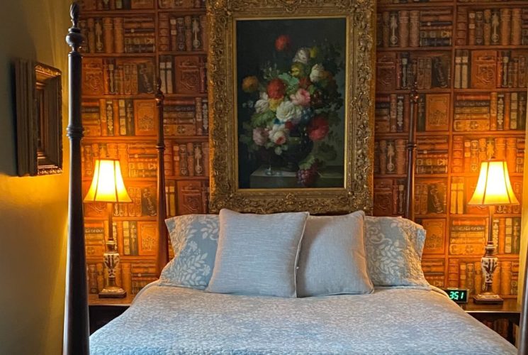 Bedroom with four poster bed against a wall covered in wallpaper that looks like bookshelves, two tables with lamps