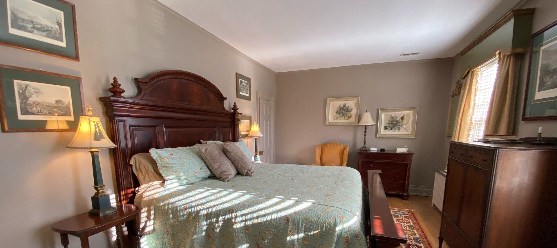 Spacious and elegant bedroom with a king bed, mahogany headboard, two dressers, chair and art work on the walls