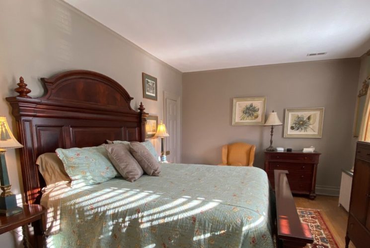 Spacious and elegant bedroom with a king bed, mahogany headboard, two dressers, chair and art work on the walls