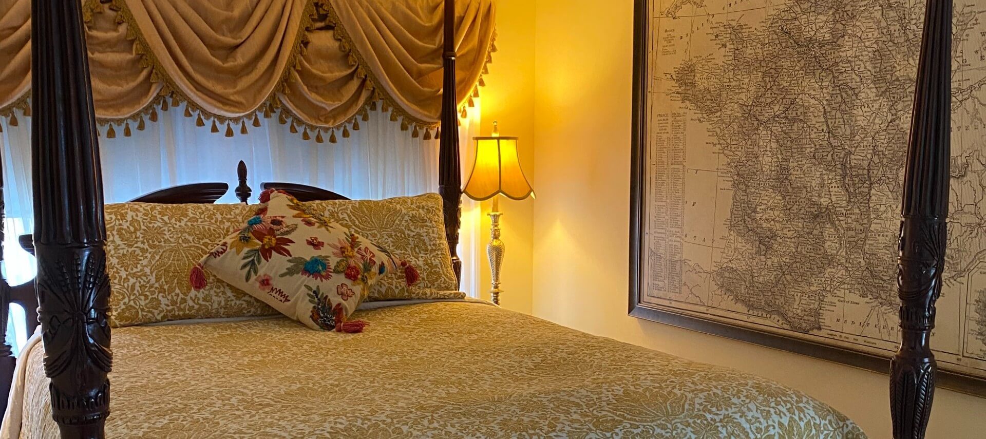 Elegant bedroom with a mohogany four poster bed, side table with lam[ and large print of a map on the wall