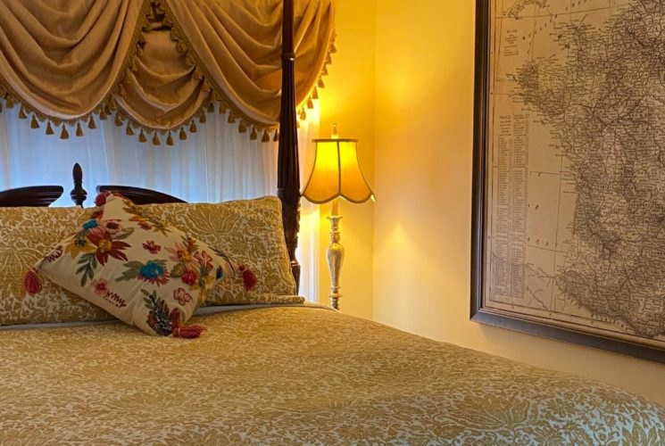 Elegant bedroom with a mohogany four poster bed, side table with lam[ and large print of a map on the wall