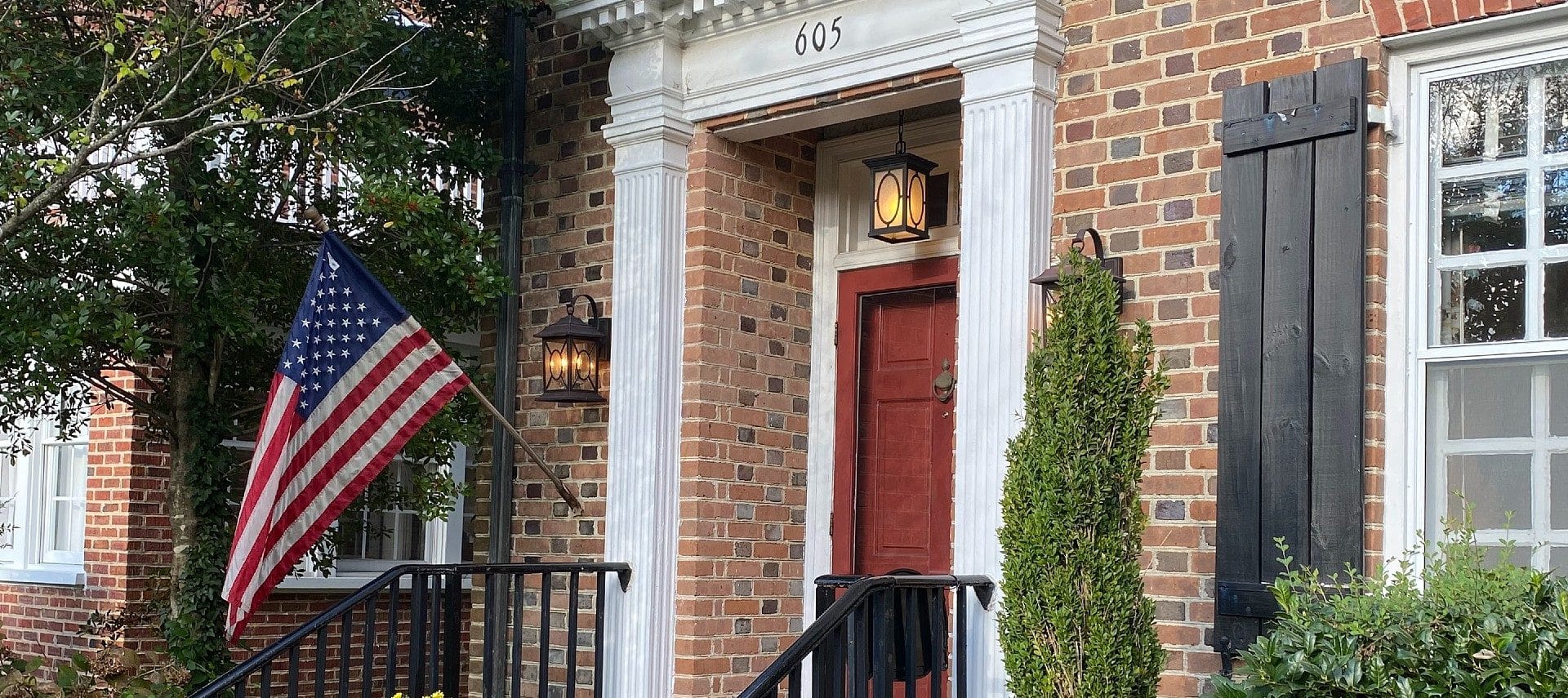 Exterior of a brick home with a red front door, black railing and shutters and an American flag on a pole