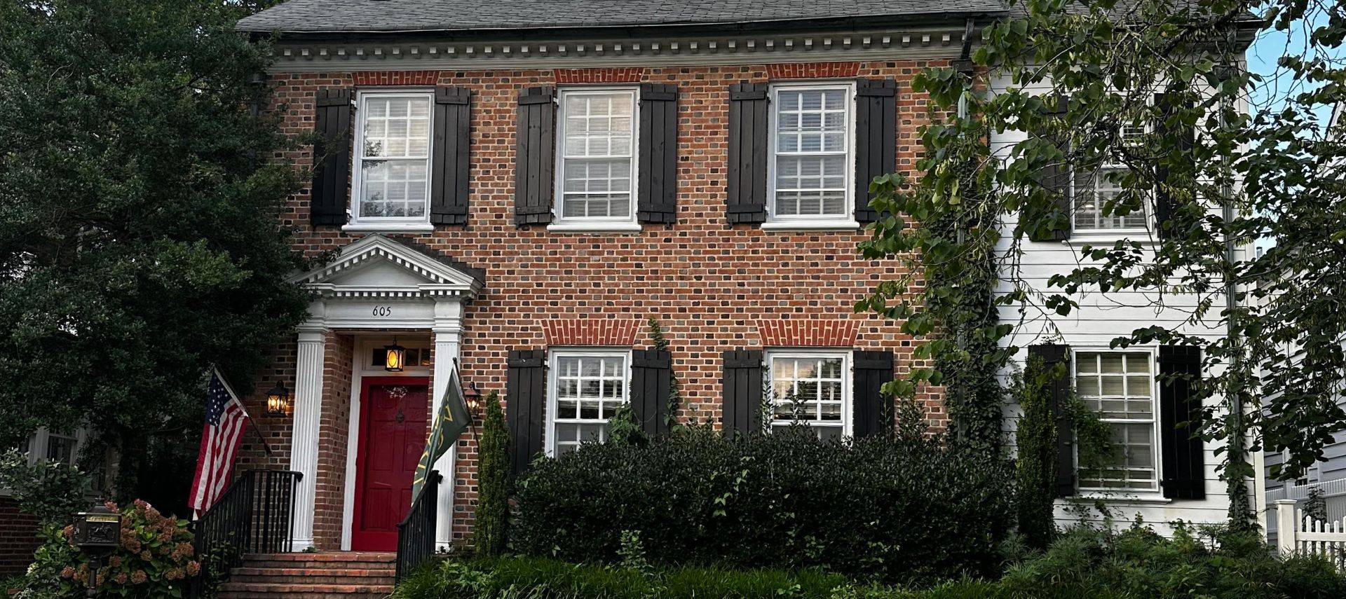 Large brick home with many windows with black shutters, red front door with the American flag on a pole