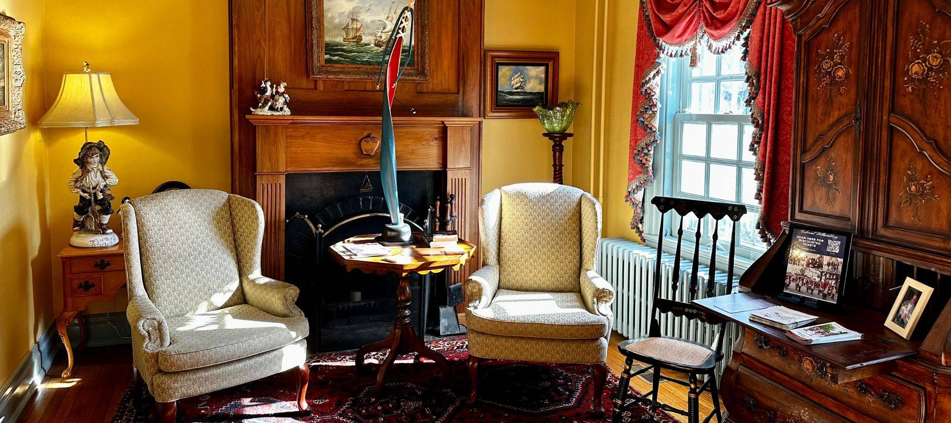 Elegant living room of a home with two wingback chairs and table in front of a fireplace, window with red curtains and tall antique hutch