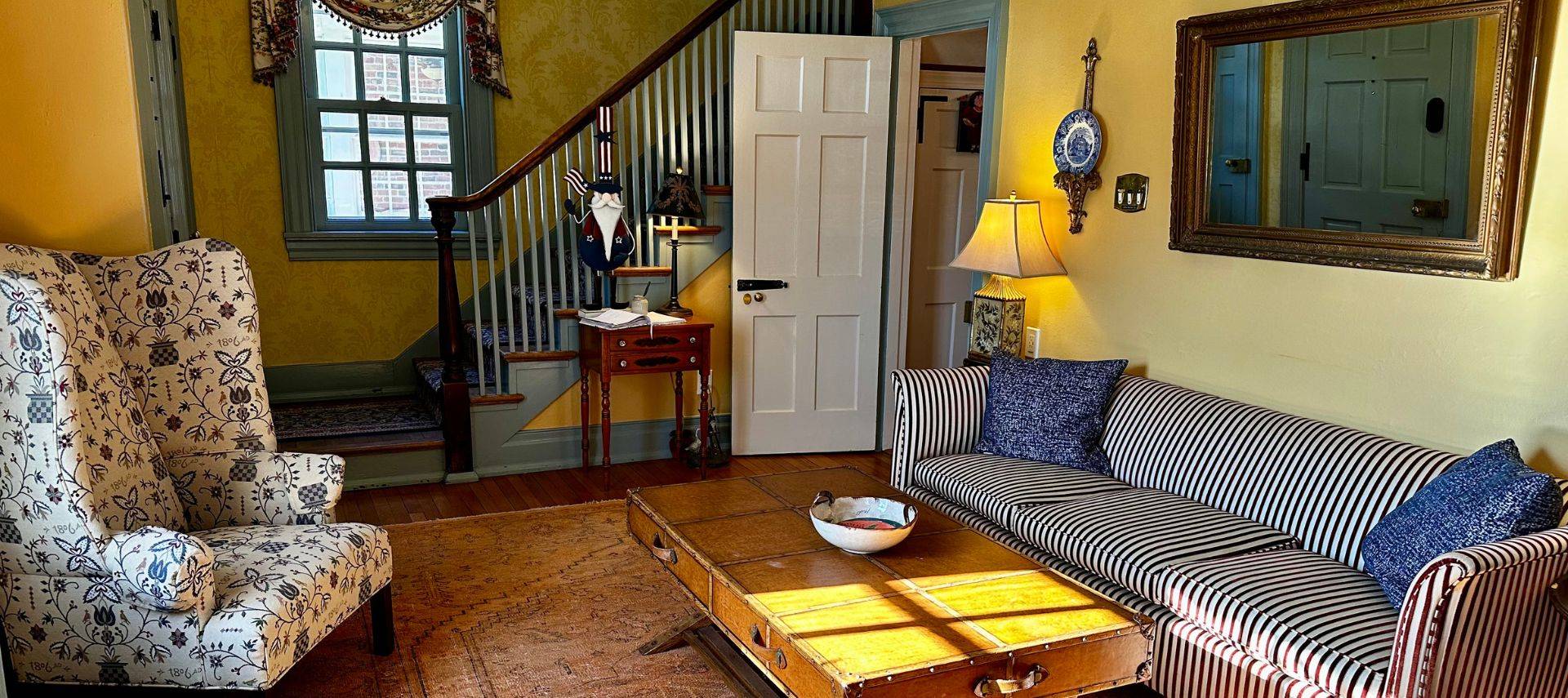 Living room with a striped couch, floral wingback chair, coffee table and stairwell to an upper level